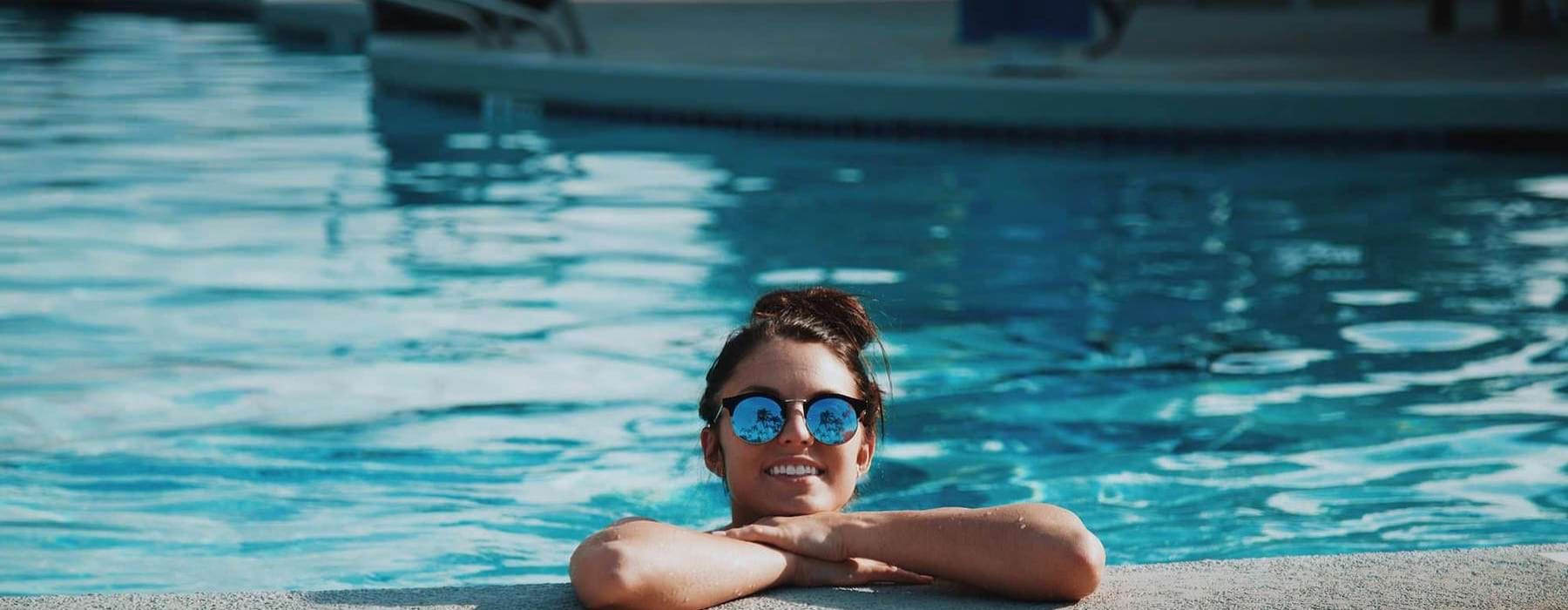 woman with sunglasses, in a pool, rests her arms on the ledge and smiles